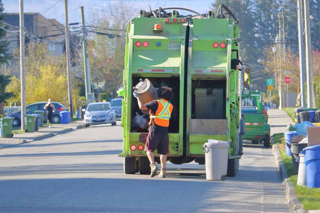 Worker unloads waste bin into collection truck on a residential street.